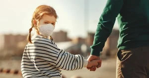 A girl wearing a mask and striped jumper is holding her dad's hand while walking to see her mom as part of a family law shared parenting arrangement during COVID 19.
