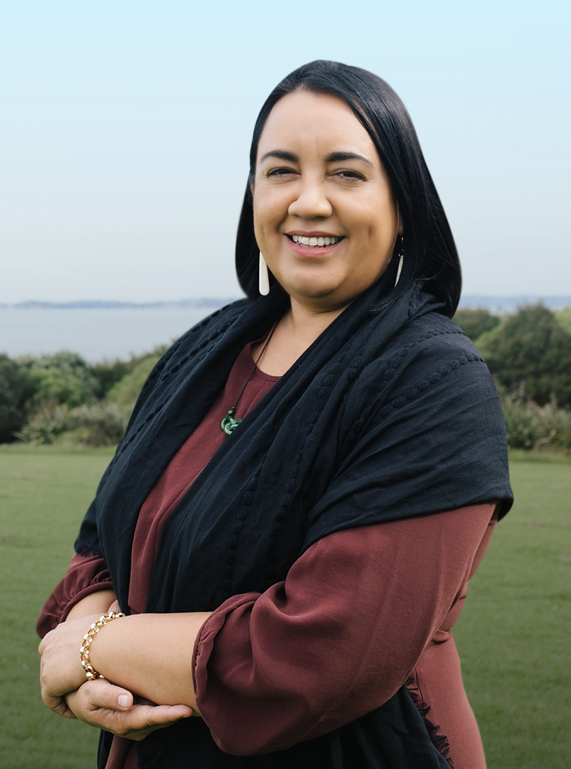 Māori Legal Lawyer Kelly Dixon, Partner of Dixon & Co Lawyers standing in the park, smiling, her holding hands together, wearing a burgundy dress with a Shaul, pounamu and while long earrings, with bushes, sea, and blue-sky background.
